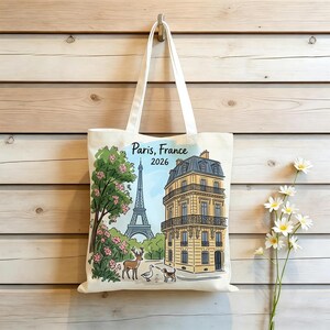 May include: A cream-colored canvas tote bag featuring a colorful illustration of the Eiffel Tower, a Parisian building, a deer, a goose, and a dog. The text "Paris, France 2026" is printed at the top. The bag has two long white fabric handles.