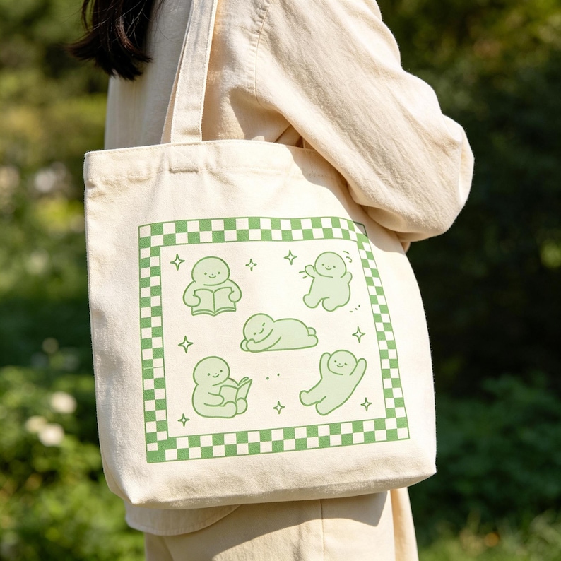 May include: Cream tote bag with a green and white chequered border. The bag features a green illustration of small figures in various poses, including reading and waving. The bag has two cream-coloured handles.