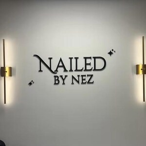 May include: A wall with the text "NAILED BY NEZ" in black lettering, flanked by two gold-colored, vertical wall sconces with integrated lighting. Small star-shaped accents are placed near the text.