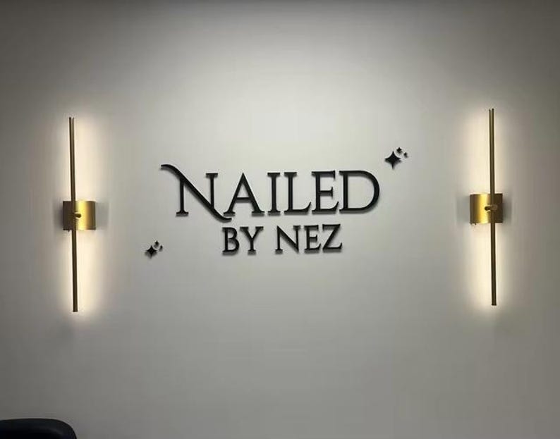 May include: A wall with the text "NAILED BY NEZ" in black lettering, flanked by two gold-colored, vertical wall sconces with integrated lighting. Small star-shaped accents are placed near the text.