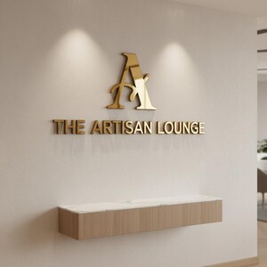 May include: Gold-colored logo and text on a white wall. The logo is a stylized letter "A" above the words "THE ARTISAN LOUNGE". Below, a wooden cabinet with a white marble top is mounted on the wall.