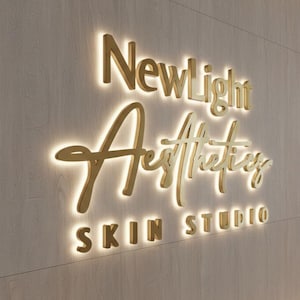 Peut inclure: Un enseigne dorée et éclairée sur un mur texturé. L'enseigne indique "Newlight Aesthetics Skin Studio" dans une police stylisée. Les lettres sont en trois dimensions et dégagent une lueur chaleureuse, créant une esthétique moderne et élégante.