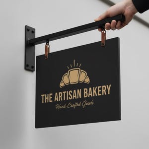 May include: A black rectangular sign for "THE ARTISAN BAKERY" with a gold croissant graphic and text. The sign is suspended by leather straps from a black metal bracket. The sign is held by a person's hand.