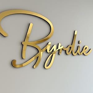 May include: A gold-colored, cursive-style sign that reads "Byrdie" is mounted on a light gray wall. The sign has a reflective, metallic finish with a black outline, creating a 3D effect. The elegant design is suitable for interior decor.