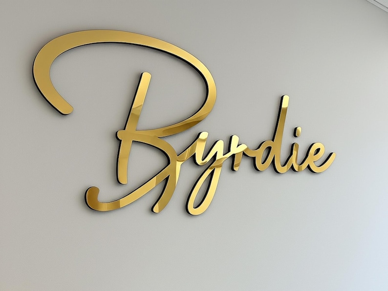 May include: A gold-colored, cursive-style sign that reads "Byrdie" is mounted on a light gray wall. The sign has a reflective, metallic finish with a black outline, creating a 3D effect. The elegant design is suitable for interior decor.