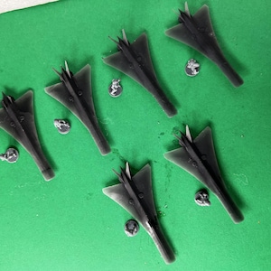 May include: Six dark gray model airplanes with a triangular wing design are arranged on a green surface. Small, circular figurines are placed near each plane. The planes have a sleek, modern aesthetic.