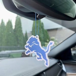 Puede incluir: Un ambientador para coche azul y blanco con forma de león, colgado de un espejo retrovisor. El león es un logotipo de equipo deportivo. El fondo muestra el interior de un coche y una vista de árboles.