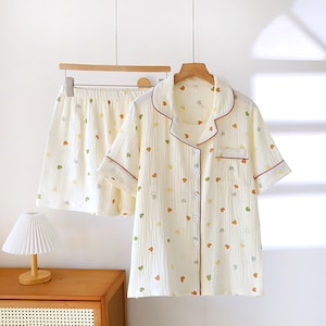 Puede incluir: Conjunto de pijama color crema con estampado de corazones. La camisa de manga corta con botones y los pantalones cortos tienen un ribete rojo y cuelgan de una percha de madera. Los corazones son de color verde, amarillo y naranja.