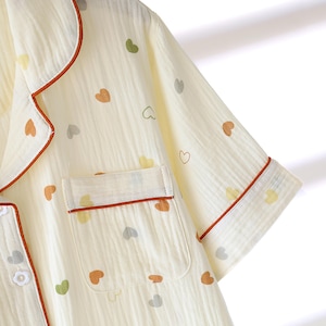 Puede incluir: Conjunto de pijama color crema con un estampado de corazones en varios colores. La parte superior de manga corta tiene un bolsillo en el pecho y est&aacute; ribeteada con una tuber&iacute;a color &oacute;xido. La tela parece ser un material ligero y texturizado.