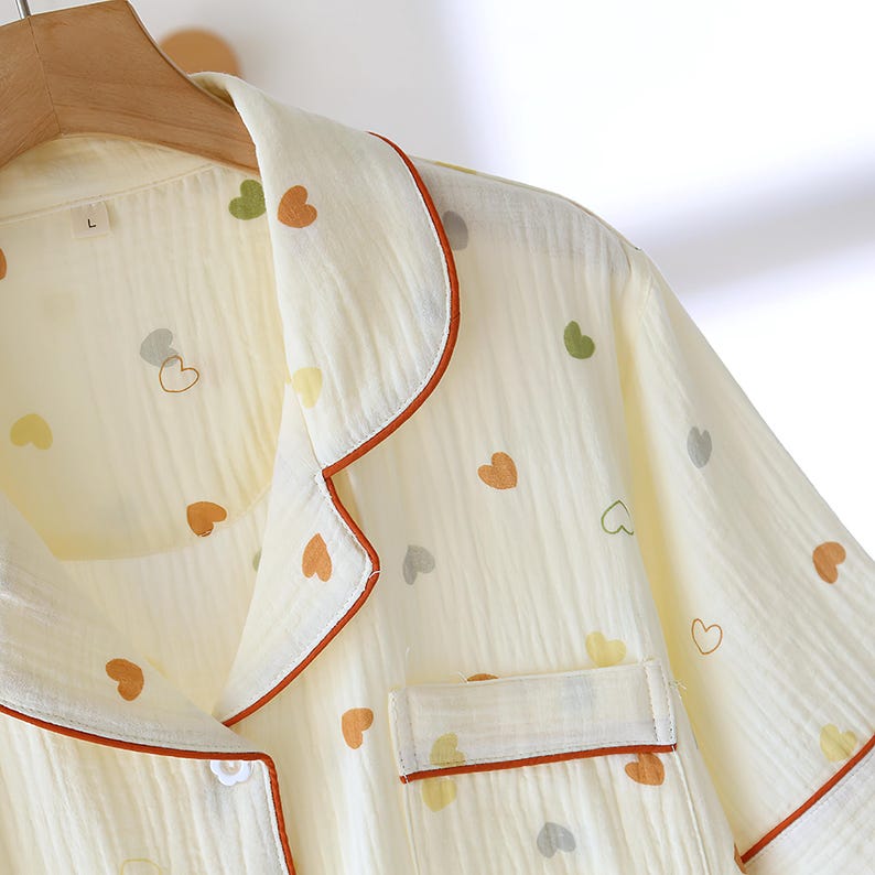 Puede incluir: Top de pijama color crema con un estampado de corazones en varios colores. La parte superior tiene un cuello con muescas y un bolsillo en el pecho, ambos ribeteados con un ribete marr&oacute;n rojizo. La tela parece ligera y texturizada, con cierre de bot&oacute;n.