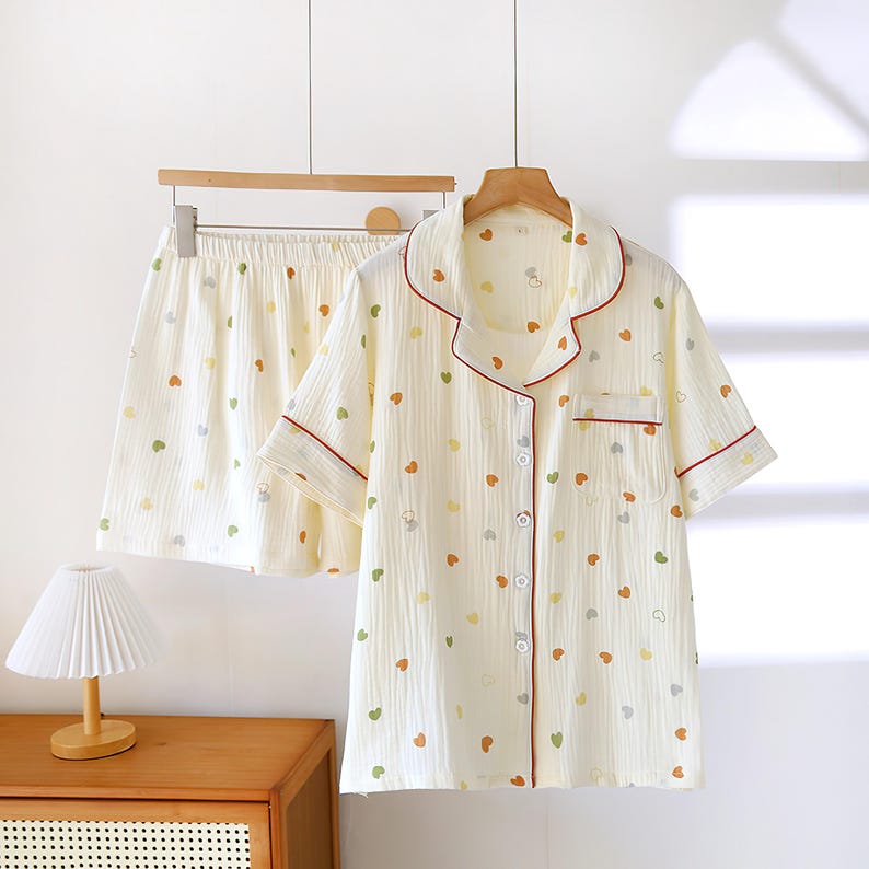 Puede incluir: Conjunto de pijama color crema con estampado de corazones. La camisa de manga corta con botones y los pantalones cortos tienen un ribete rojo y cuelgan de una percha de madera. Los corazones son de color verde, amarillo y naranja.