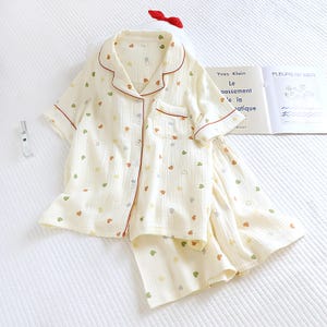 Puede incluir: Conjunto de pijama color crema con un estampado de corazones multicolores. La parte superior de manga corta y los pantalones cortos tienen ribetes rojos y cierres de botones. Tambi&eacute;n se ve un lazo rojo y un libro.