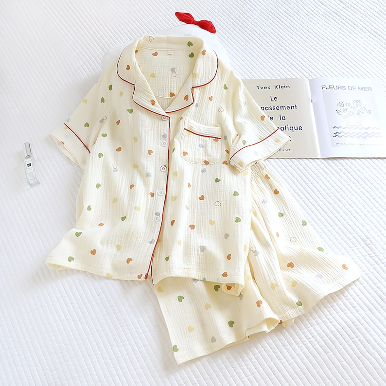 Puede incluir: Conjunto de pijama color crema con un estampado de corazones multicolores. La parte superior de manga corta y los pantalones cortos tienen ribetes rojos y cierres de botones. Tambi&eacute;n se ve un lazo rojo y un libro.