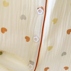 Puede incluir: Primer plano de una parte superior de pijama color crema con un estampado de corazones en tonos marr&oacute;n, gris y amarillo. La parte superior presenta botones en forma de flor y un ribete color &oacute;xido a lo largo de la l&iacute;nea de botones.
