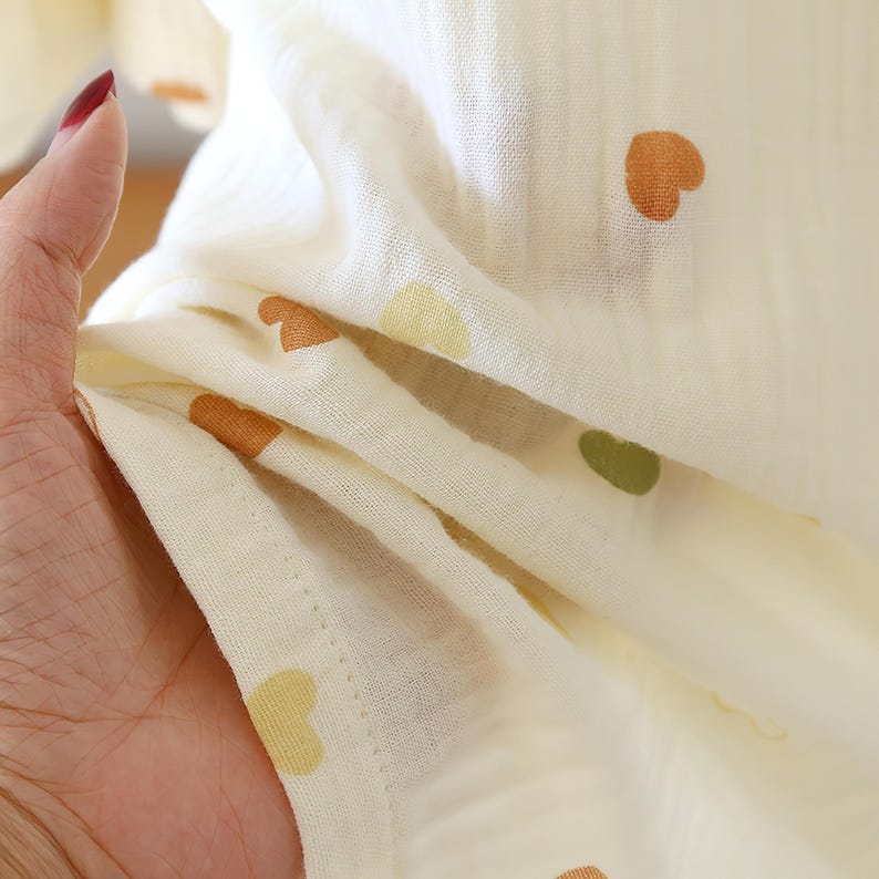 Puede incluir: Primer plano de una tela color crema con un estampado de corazones. Los corazones son en tonos marr&oacute;n, amarillo y verde. La tela es suave y parece ligera, con pliegues y texturas visibles.