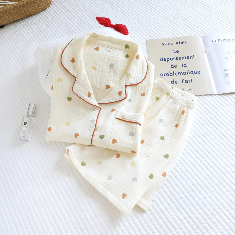 Puede incluir: Conjunto de pijama color crema con un estampado de corazones en varios colores. El conjunto incluye una camisa abotonada con ribete rojo y pantalones a juego. Tambi&eacute;n se ven un lazo rojo y un libro con el texto "Yves Klein".