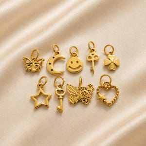 May include: A collection of nine gold-toned metal charms featuring various shapes including a butterfly, a crescent moon with stars, a smiley face, keys, a four-leaf clover, a star outline, a detailed butterfly, and a beaded heart with a star.