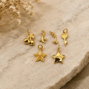 May include: A collection of six gold-toned metal charms featuring a pair of cherries, an airplane, an umbrella, a heart-topped key, a five-pointed star, and a five-pointed star with the word Lucky embossed on the front, each with a jump ring.