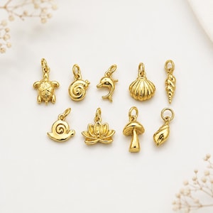 Puede incluir: Una colección de nueve pequeños dijes de metal en tono dorado con diversas formas, incluyendo una tortuga, una concha en espiral, un delfín, una concha de abanico, una concha alargada, un caracol, una flor de loto, un hongo y una pequeña concha.