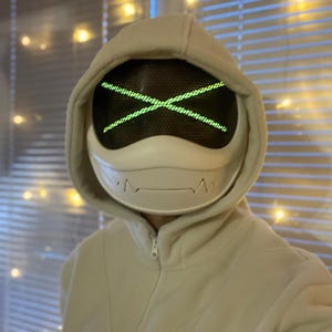 Peut inclure: Un masque blanc futuriste avec un écran en maille et un motif de croix verte lumineuse. Le masque fait partie d'un sweat à capuche, créant un look moderne et inspiré de la technologie. La palette de couleurs est neutre, avec l'accent mis sur le design unique du masque.