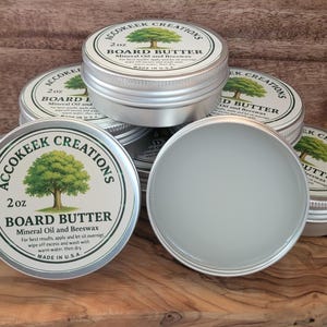 May include: Several small, silver-colored tins of board butter are stacked on a wooden cutting board. The tins have white labels with a green tree logo and the text "BOARD BUTTER". One tin is open, revealing the product inside. The background is a wooden surface.