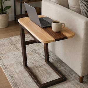 May include: A C-shaped side table featuring a two-tone wooden top with light and dark wood grain panels. The base is constructed from dark brown wood, forming a sturdy frame that slides under furniture. A laptop and a ceramic mug sit on top.