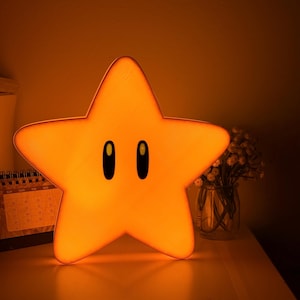 K&ouml;nnte beinhalten: Ein sternf&ouml;rmiges Nachtlicht, das ein warmes orangefarbenes Leuchten ausstrahlt. Die Lampe hat ein Cartoon-Design mit zwei schwarzen ovalen Augen. Sie steht auf einer wei&szlig;en Oberfl&auml;che.