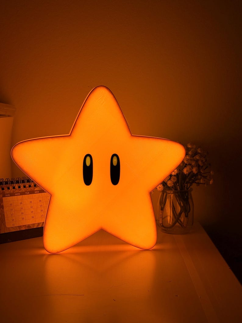 K&ouml;nnte beinhalten: Ein sternf&ouml;rmiges Nachtlicht, das ein warmes orangefarbenes Leuchten ausstrahlt. Die Lampe hat ein Cartoon-Design mit zwei schwarzen ovalen Augen. Sie steht auf einer wei&szlig;en Oberfl&auml;che.