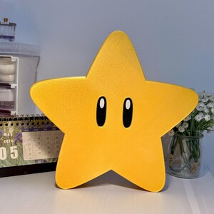 K&ouml;nnte beinhalten: Eine gro&szlig;e, gelbe sternf&ouml;rmige Lampe mit schwarzen, ovalen Augen. Die Lampe hat eine leuchtend gelbe Farbe und eine glatte Oberfl&auml;che. Der Stern ist ein cartoonartiges Design, das an eine Videospiel-Figur erinnert. Die Lampe steht auf einer wei&szlig;en Oberfl&auml;che.