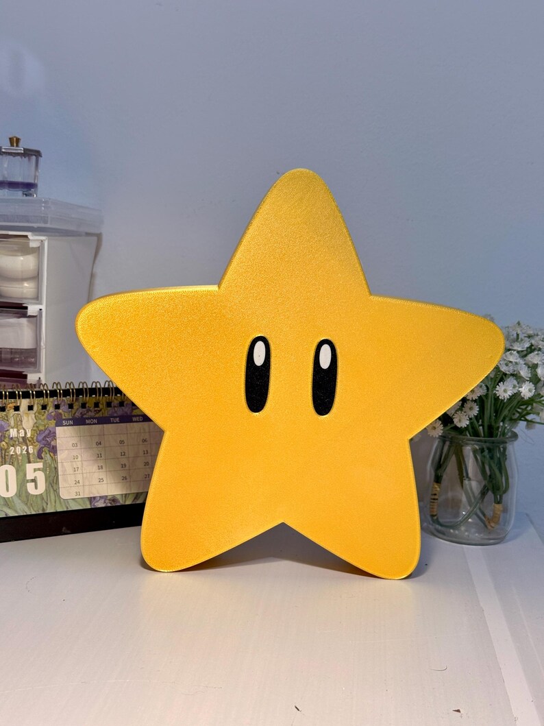 K&ouml;nnte beinhalten: Eine gro&szlig;e, gelbe sternf&ouml;rmige Lampe mit schwarzen, ovalen Augen. Die Lampe hat eine leuchtend gelbe Farbe und eine glatte Oberfl&auml;che. Der Stern ist ein cartoonartiges Design, das an eine Videospiel-Figur erinnert. Die Lampe steht auf einer wei&szlig;en Oberfl&auml;che.