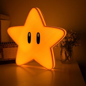 K&ouml;nnte beinhalten: Eine leuchtend gelbe sternf&ouml;rmige Nachtlampe mit schwarzen ovalen Augen. Das Licht steht auf einer wei&szlig;en Oberfl&auml;che, im Hintergrund ein Kalender und eine Vase mit Blumen. Der Stern ist eine beliebte Figur aus einem Videospiel.