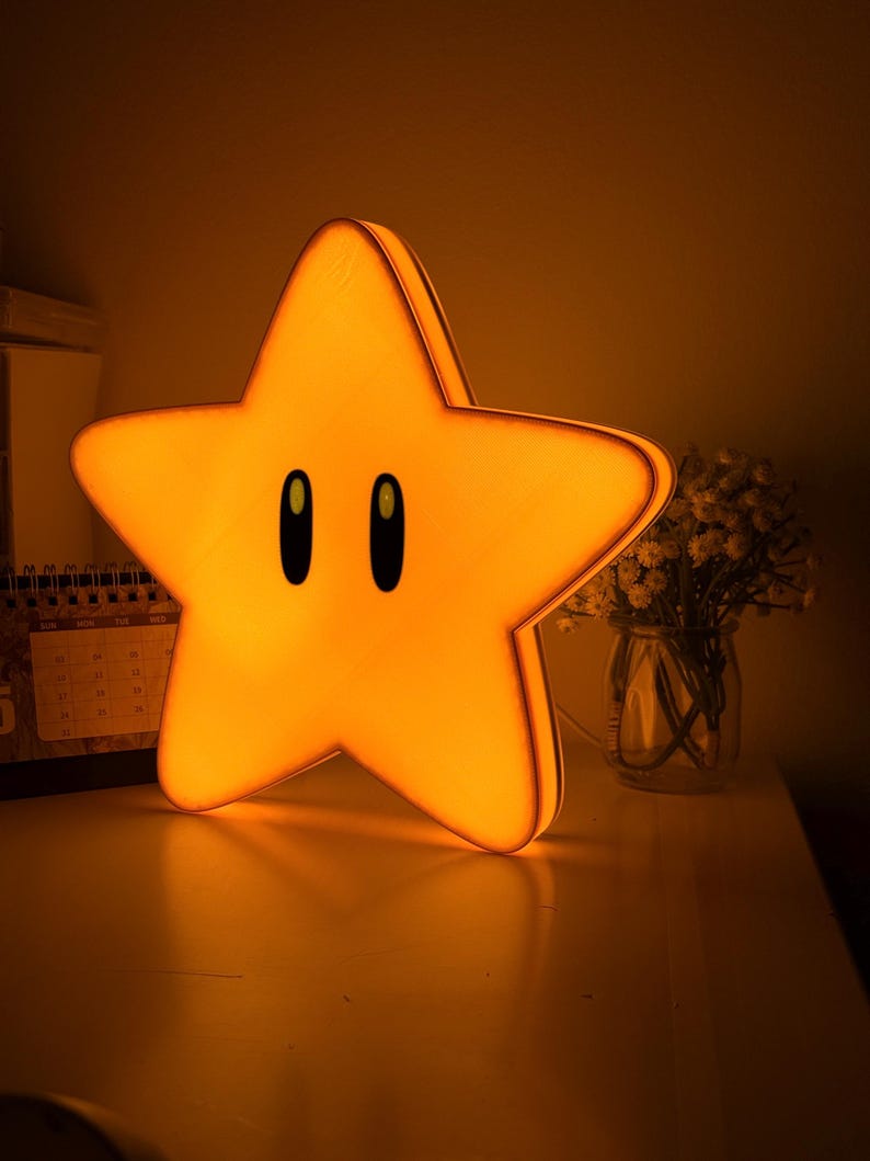 K&ouml;nnte beinhalten: Eine leuchtend gelbe sternf&ouml;rmige Nachtlampe mit schwarzen ovalen Augen. Das Licht steht auf einer wei&szlig;en Oberfl&auml;che, im Hintergrund ein Kalender und eine Vase mit Blumen. Der Stern ist eine beliebte Figur aus einem Videospiel.