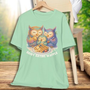 Puede incluir: Una camiseta verde claro con una colorida ilustraci&oacute;n de tres b&uacute;hos disfrutando de gofres. Los b&uacute;hos tienen plumas vibrantes y est&aacute;n reunidos alrededor de un plato de gofres, pl&aacute;tanos y bayas. El texto "ENJOY EATING WAFFLE" est&aacute; impreso debajo.