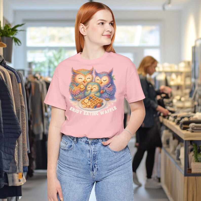 Puede incluir: Camiseta rosa claro con una colorida ilustraci&oacute;n de tres b&uacute;hos disfrutando de gofres. Los b&uacute;hos est&aacute;n representados en tonos naranja, azul y verde. El texto "ENJOY EATING WAFFLE" est&aacute; impreso debajo.