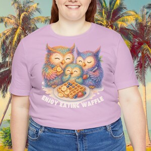 Puede incluir: Una camiseta lila claro con una ilustraci&oacute;n colorida de tres b&uacute;hos comiendo gofres. Los b&uacute;hos est&aacute;n representados en colores brillantes, con el texto "ENJOY EATING WAFFLE" debajo de la imagen. La camiseta es de material suave.