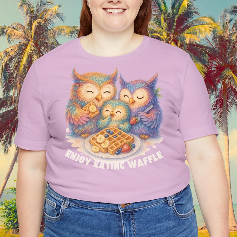 Puede incluir: Una camiseta lila claro con una ilustraci&oacute;n colorida de tres b&uacute;hos comiendo gofres. Los b&uacute;hos est&aacute;n representados en colores brillantes, con el texto "ENJOY EATING WAFFLE" debajo de la imagen. La camiseta es de material suave.