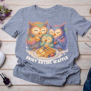 Puede incluir: Camiseta gris claro con una colorida ilustraci&oacute;n de tres b&uacute;hos comiendo gofres con pl&aacute;tanos y ar&aacute;ndanos. El texto "ENJOY EATING WAFFLE" est&aacute; impreso debajo.
