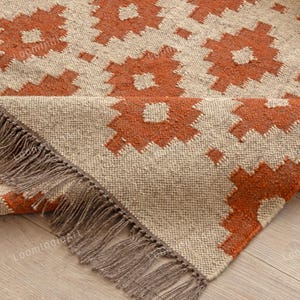 以下が含まれることがあります： オレンジとベージュの色合いの幾何学模様の織りラグ。 ラグはフリンジエッジで、天然繊維素材で作られています。 デザインは、繰り返される花のような形が特徴です。
