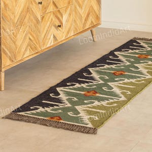 Op de afbeelding: Een lange, rechthoekige loper met een geometrisch patroon in groen, zwart en beige tinten. De loper heeft een gefranjerde rand en ligt op een lichtgekleurde vloer. Het ontwerp bevat oranje accenten.