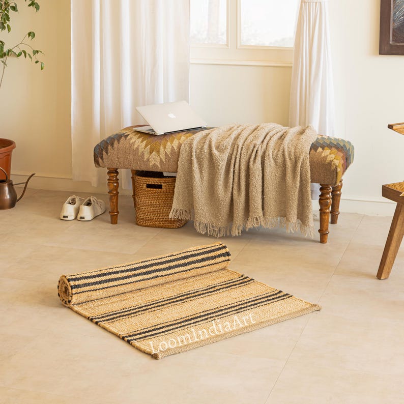 May include: A rolled-up, rectangular jute rug with black stripes is on a light-colored floor. The rug is in front of a bench with a patterned cushion and a beige blanket. A laptop sits on the bench.