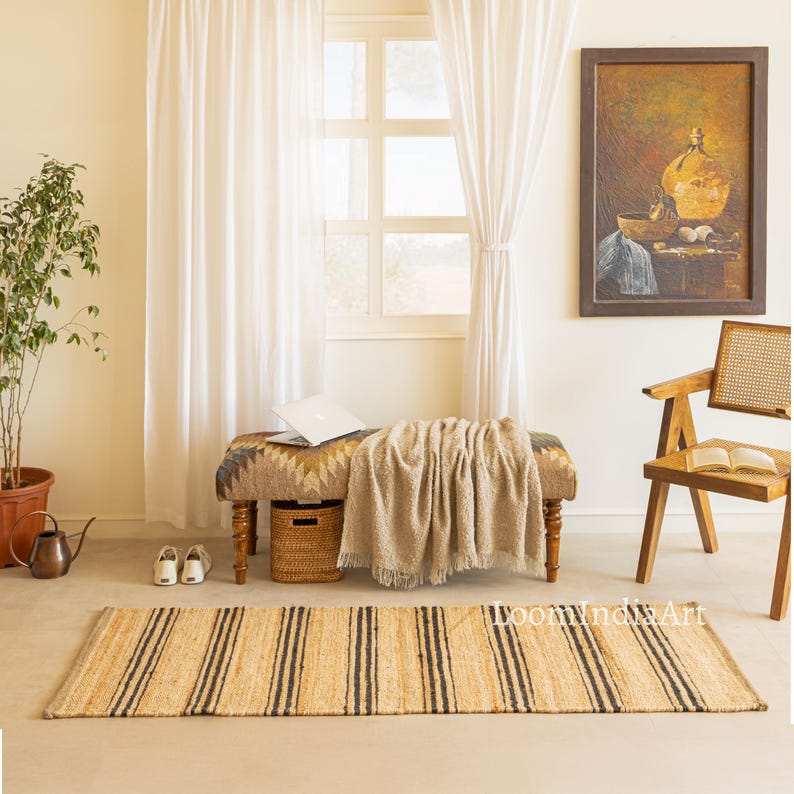 May include: A natural fiber rug with vertical black stripes. The rug is rectangular and lies on a light-colored floor. The room also features a bench, a blanket, a painting, and a window with white curtains.