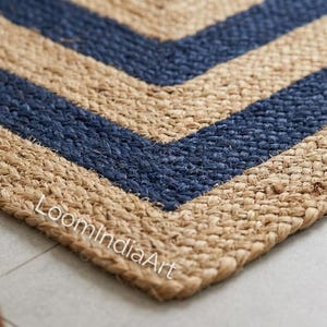 Puede incluir: Una alfombra rectangular tejida con fibras de yute de color tostado natural que presenta un llamativo patrón de chevrón azul marino. La superficie tiene una construcción trenzada gruesa y texturizada. El texto LoomIndiaArt está impreso en blanco.