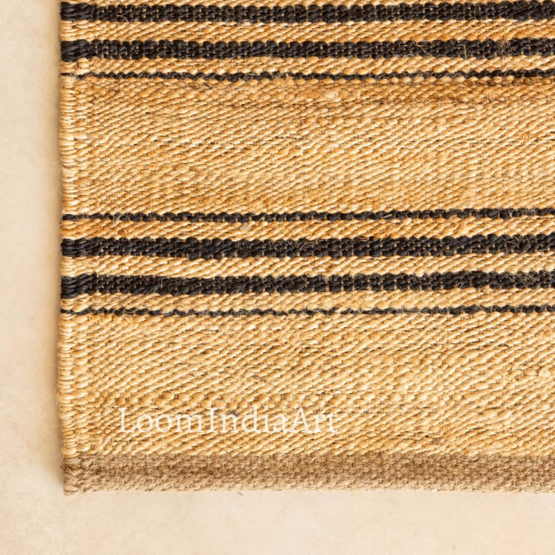 May include: Close-up of a natural fiber rug with a woven texture. The rug features horizontal black stripes against a beige background. The bottom edge has a darker brown border. The text "LoomIndiaArt" is visible.