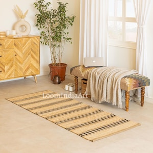 May include: A rectangular jute rug with black stripes. The rug is placed on a light-colored floor in a room with a bench, a wooden cabinet, and a potted plant. The rug has the text "LoomIndiaArt" printed on it.