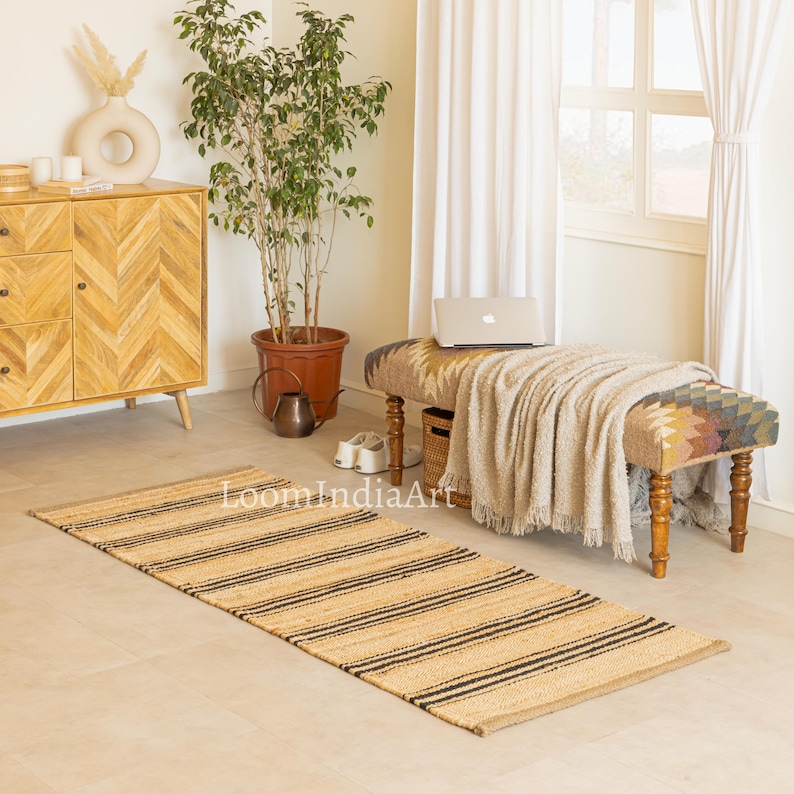 May include: A rectangular jute rug with black stripes. The rug is placed on a light-colored floor in a room with a bench, a wooden cabinet, and a potted plant. The rug has the text "LoomIndiaArt" printed on it.