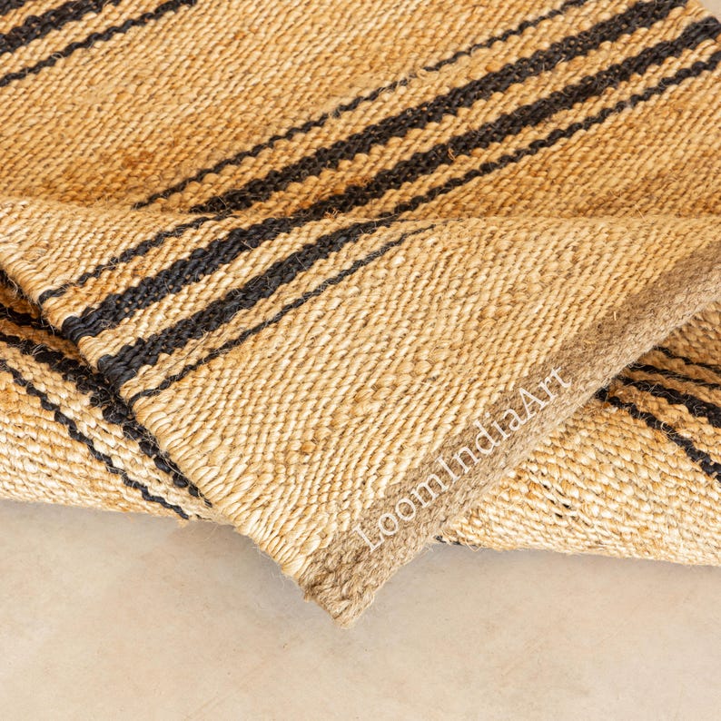 May include: A natural fiber rug with a woven texture. The rug features bold black stripes against a beige background. The edge of the rug is a lighter brown color. The text "LoomIndiaArt" is visible on the rug.