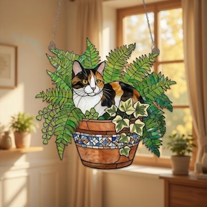 Könnte beinhalten: Hängende Dekoration aus Buntglas mit einer Katze in einem Blumentopf. Die Katze hat ein Schildpattmuster aus Schwarz, Orange und Weiß. Der Topf ist braun mit einem blau-weißen Mosaikdesign. Grüne Farne und Efeu umgeben die Katze.