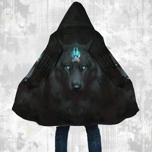 Puede incluir: Capa con capucha negra con una ilustración de lobo. El lobo tiene ojos azul brillante y un diseño de huella de pata en la frente. La capa es de tela oscura y tiene un diseño fluido.