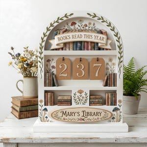 May include: A white wooden bookshelf with an arched top, decorated with floral and mushroom motifs. It features shelves with miniature books and wooden tags displaying the numbers 237. The banner reads "BOOKS READ THIS YEAR" and "Mary's Library".