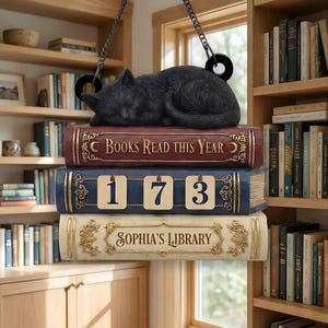 Könnte beinhalten: Ein dekoratives, hängendes Bücherregal mit drei gestapelten Büchern. Eine schwarze Katzenfigur liegt auf dem obersten Buch, auf dem "Books Read This Year" steht. Das mittlere Buch zeigt die Zahlen "173". Das untere Buch trägt die Aufschrift "Sophia's Library".